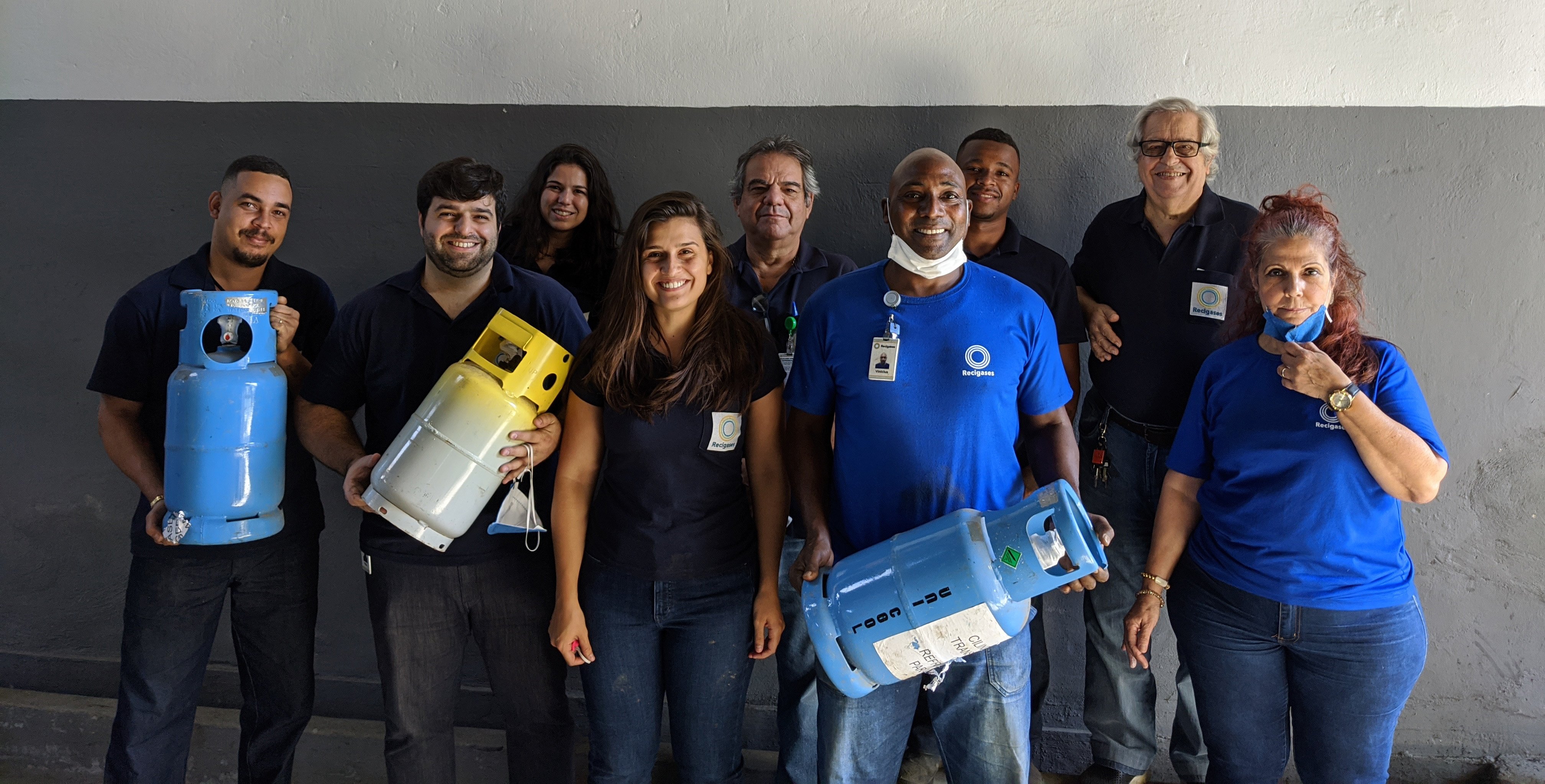 A foto está composta por 09 colaboradores olhando para a câmera e sorrindo. Dentre esses, três colaboradores estão segurando cilindros de gás refrigerantes.